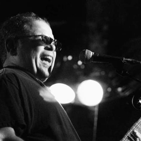 Singer in sunglasses performing at a microphone on a dimly lit stage, with bright lights behind him