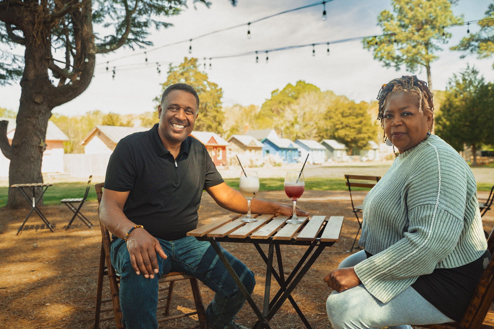 Two people seated at a patio table with drinks, smiling in an outdoor garden setting.