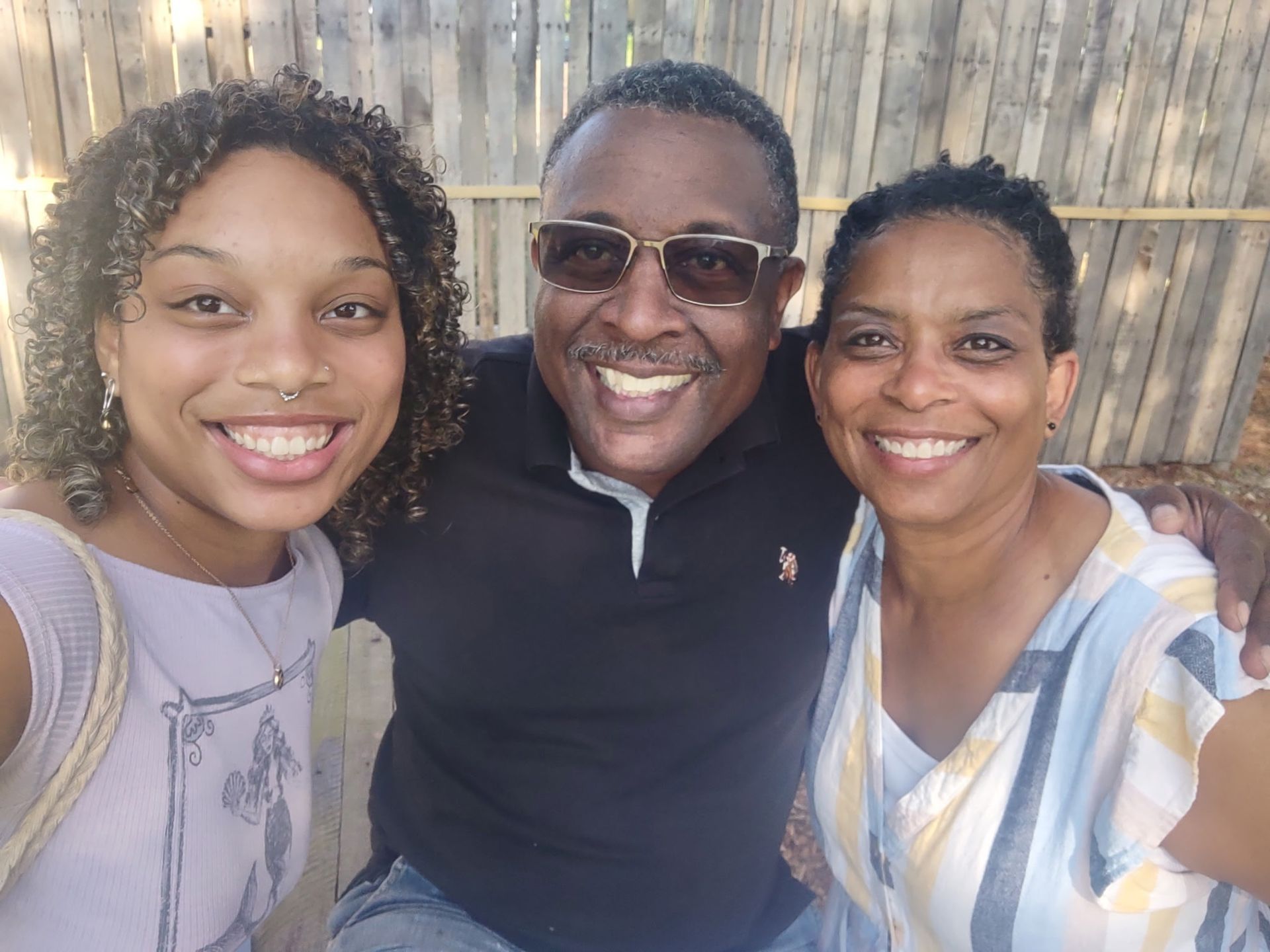 Three people smiling outdoors in front of a wooden fence, posing closely together.