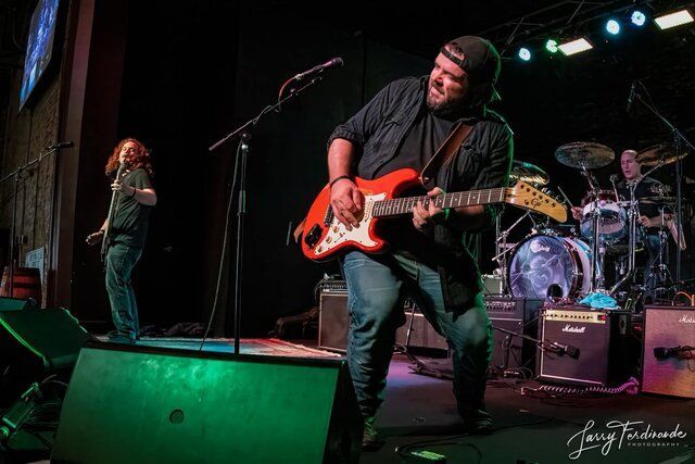 Rock band performing onstage, guitarist in black playing red electric guitar under green and purple lights.
