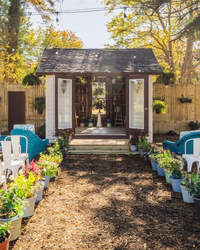 Garden path leads to a small shed with open doors, flanked by colorful potted plants and chairs.