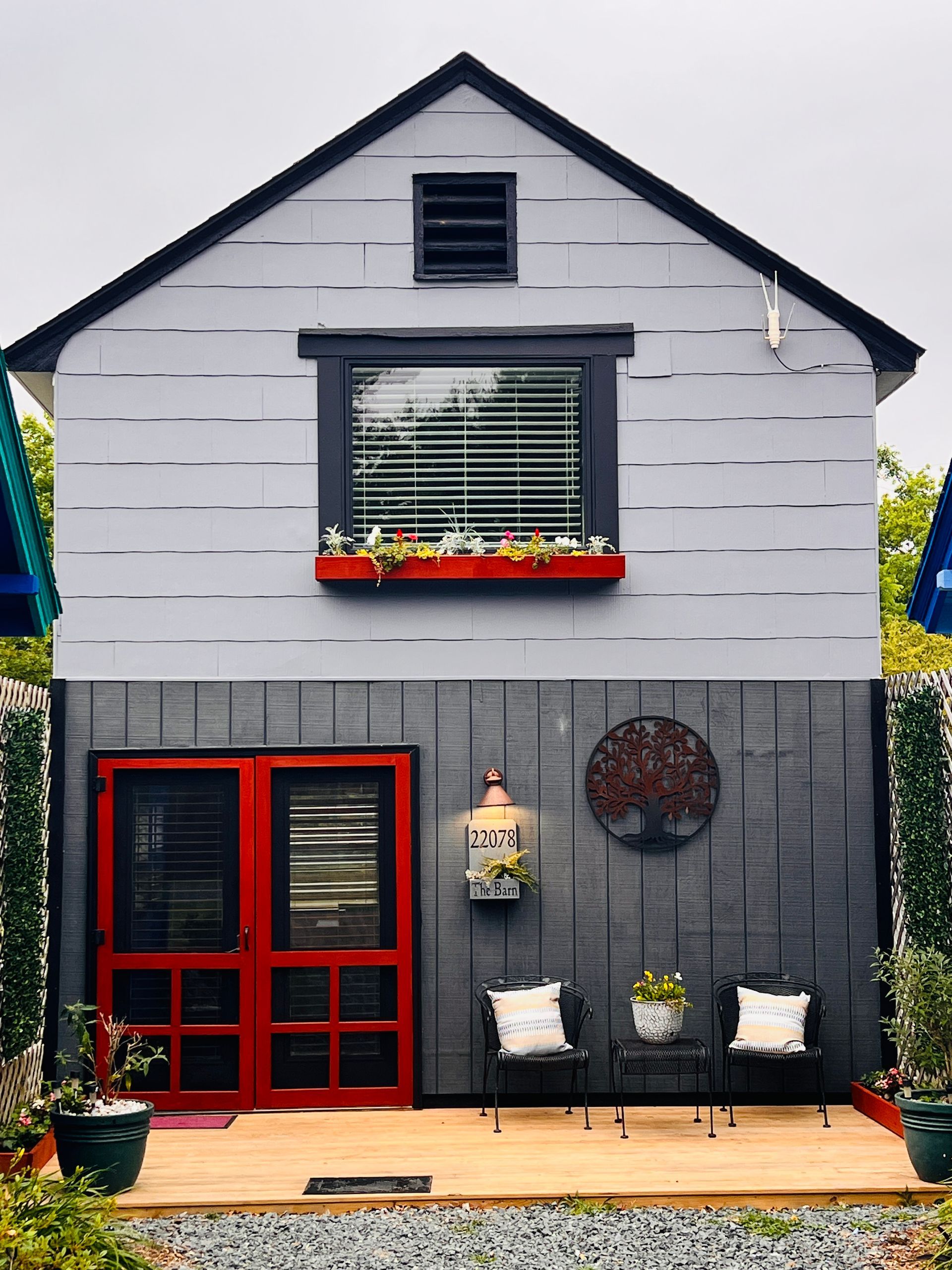 Small gray house facade with red-framed door, window boxes, and a wooden deck with outdoor chairs.