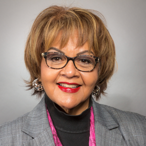 Arlene Young with glasses, smiling, wearing a gray blazer, black turtleneck, and pink scarf.