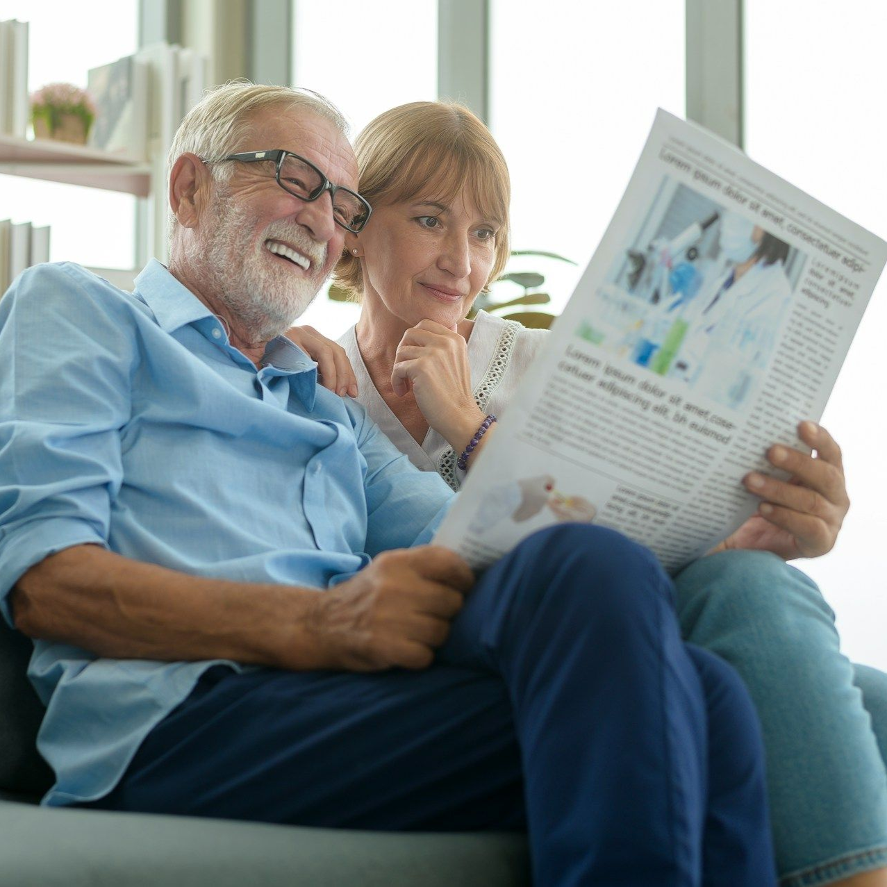 Elderly couple sitting on a couch, reading a newspaper, smiling.