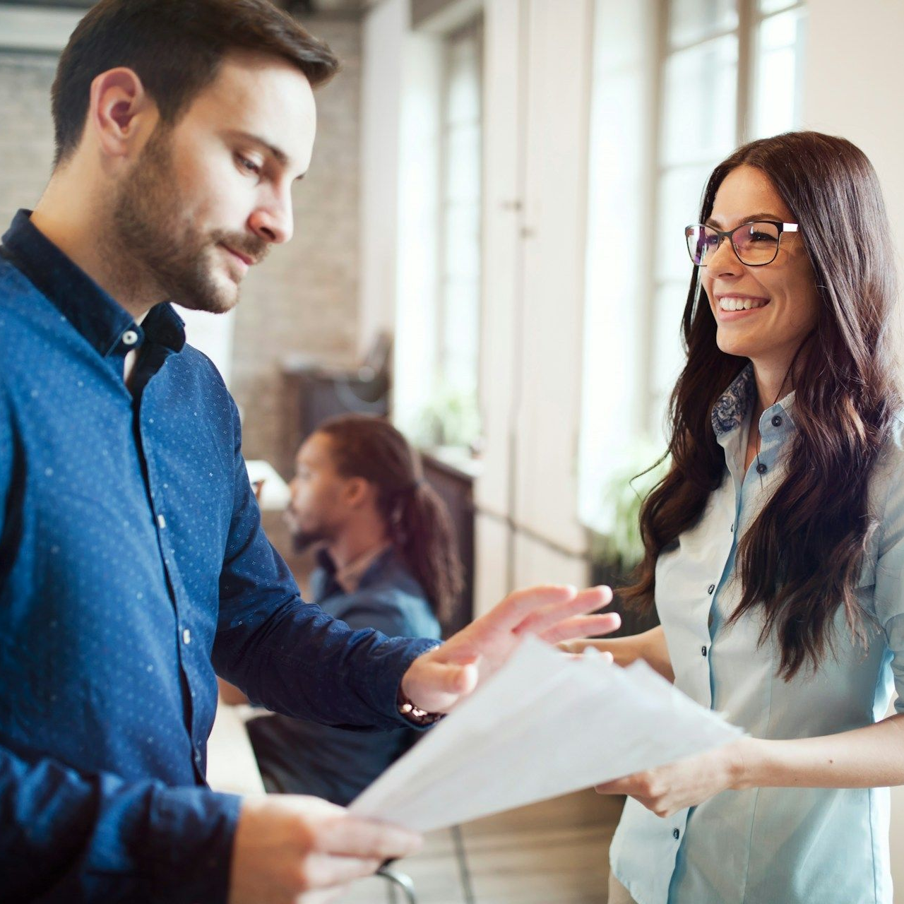 Man and woman in office, reviewing documents. Woman smiles, man gestures with papers.