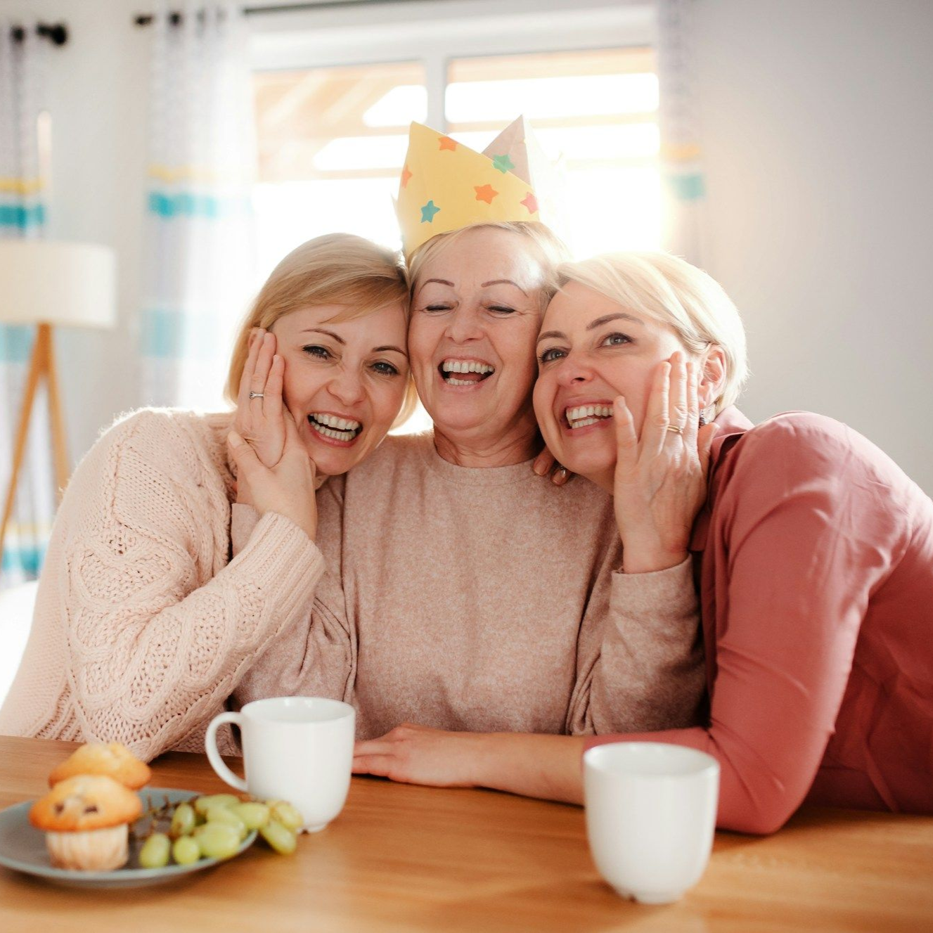 Three women laughing together, one wearing a paper crown, at a table with food and drinks.