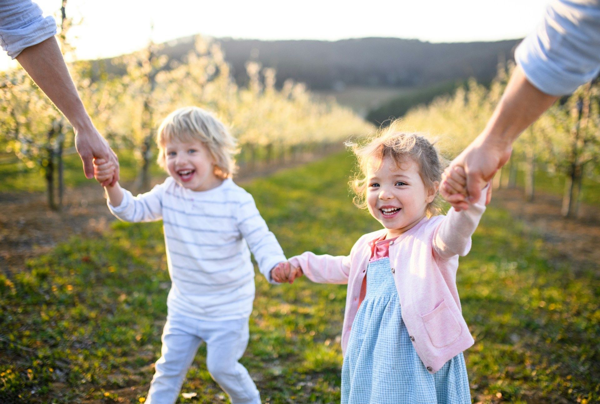 Two children smiling, holding hands with unseen adults in a sunny orchard.