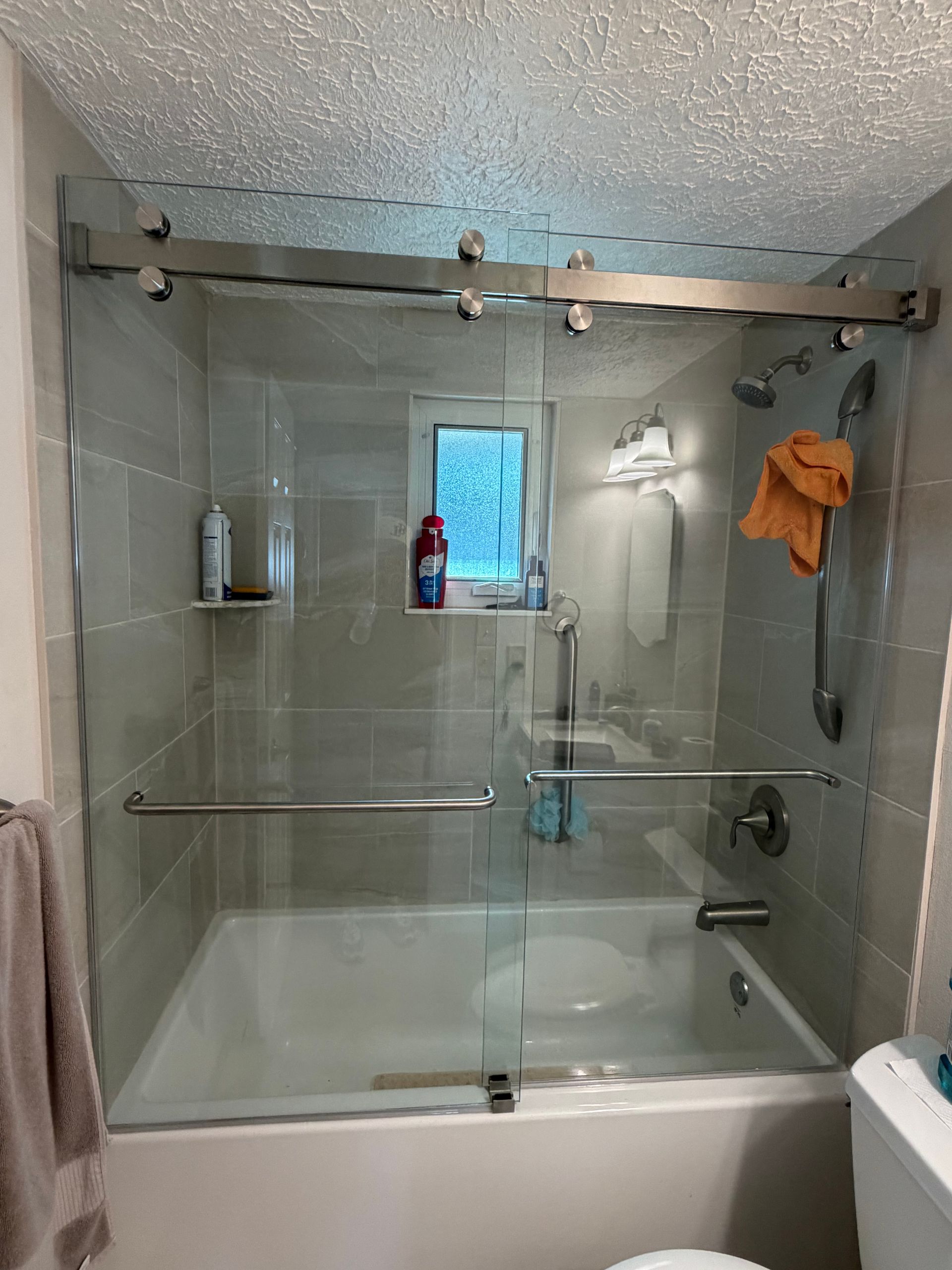 Small bathroom with a glass-enclosed bathtub shower, gray tile walls, and a window above the tub.