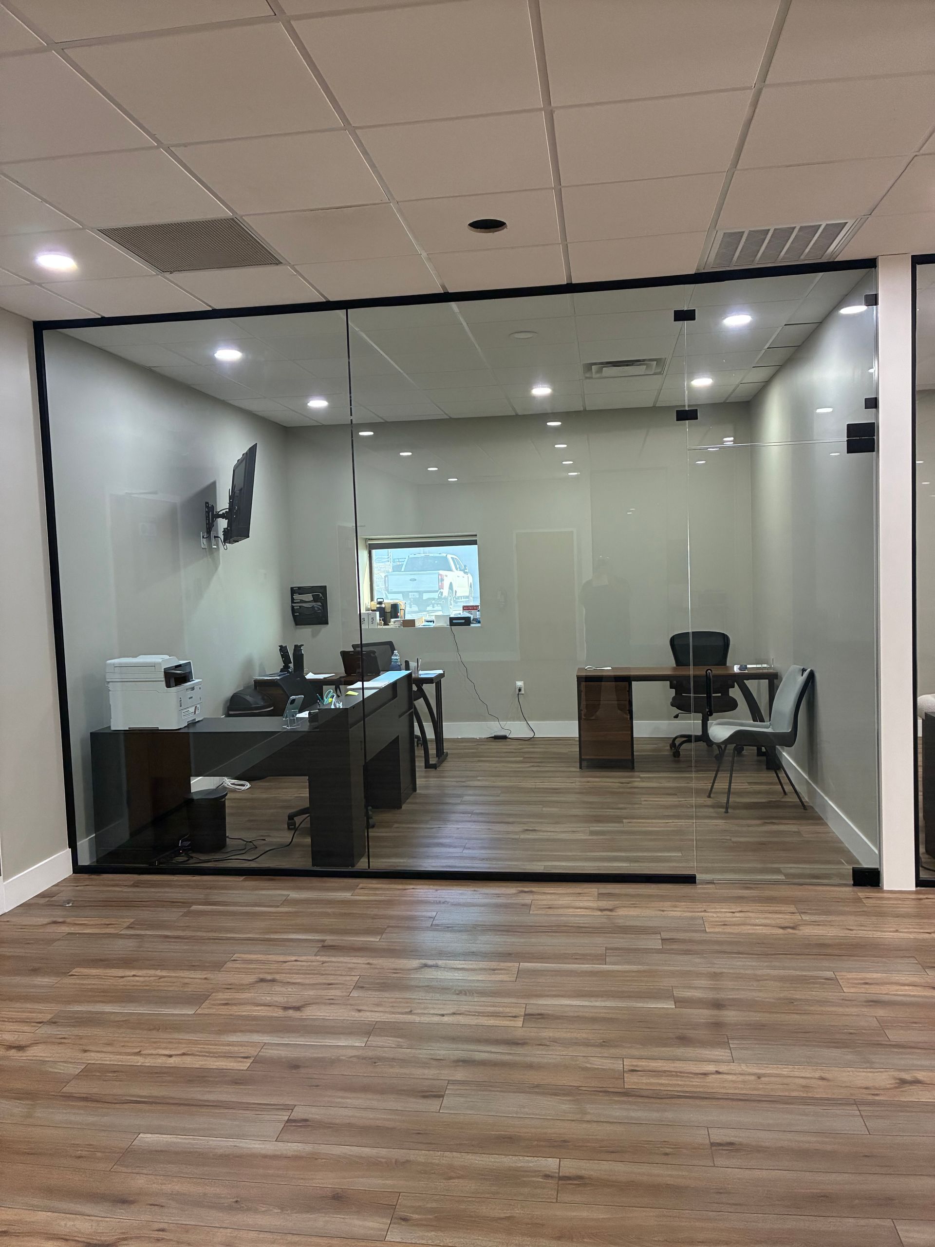 Empty glass-walled office with desks, monitors, and wood flooring inside a modern workspace