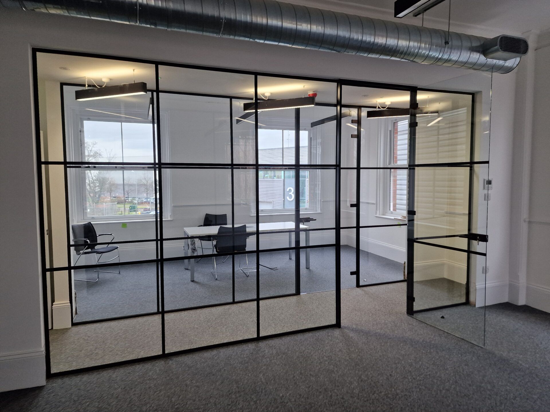 Modern industrial office interior featuring a conference room enclosed by black grid-patterned glass walls and exposed ceiling ductwork, providing a bright, open workspace division similar to frameless glass office partitions.