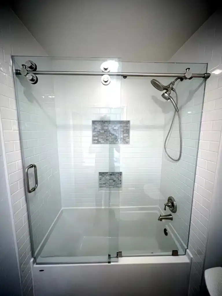 A clean, modern bathroom alcove shower featuring a sleek, frameless clear glass enclosure and a sophisticated sliding glass shower door system with visible top-mounted roller wheels. The interior is tiled with white subway tiles, and a tiled mosaic shelf niche and polished chrome shower fixtures are visible.