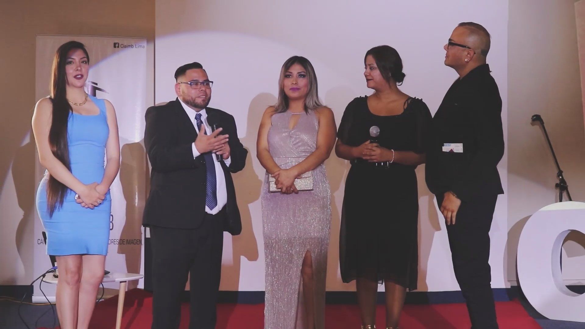 Cinco personas están de pie en un escenario, vestidas de manera formal, y un hombre habla por un micrófono.