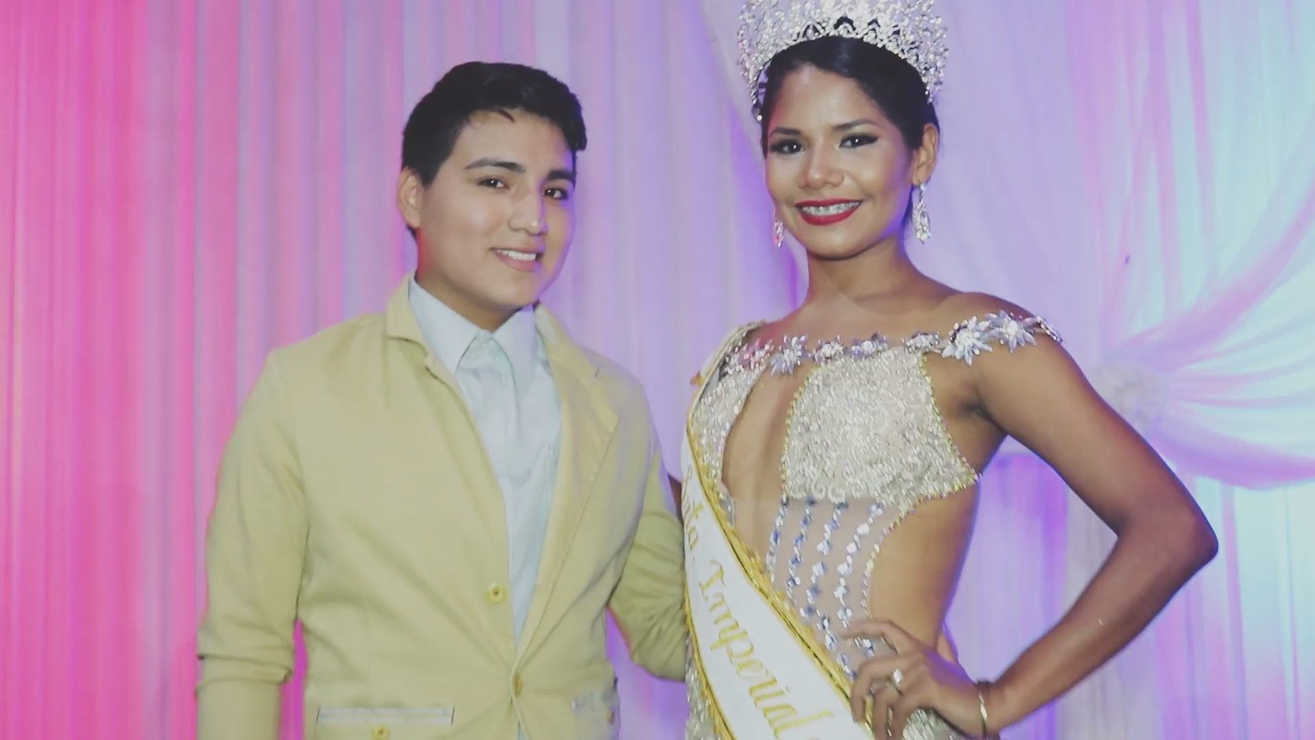 Dos personas vestidas de manera formal posan frente a un fondo rosa con cortinas, ambas sonriendo.