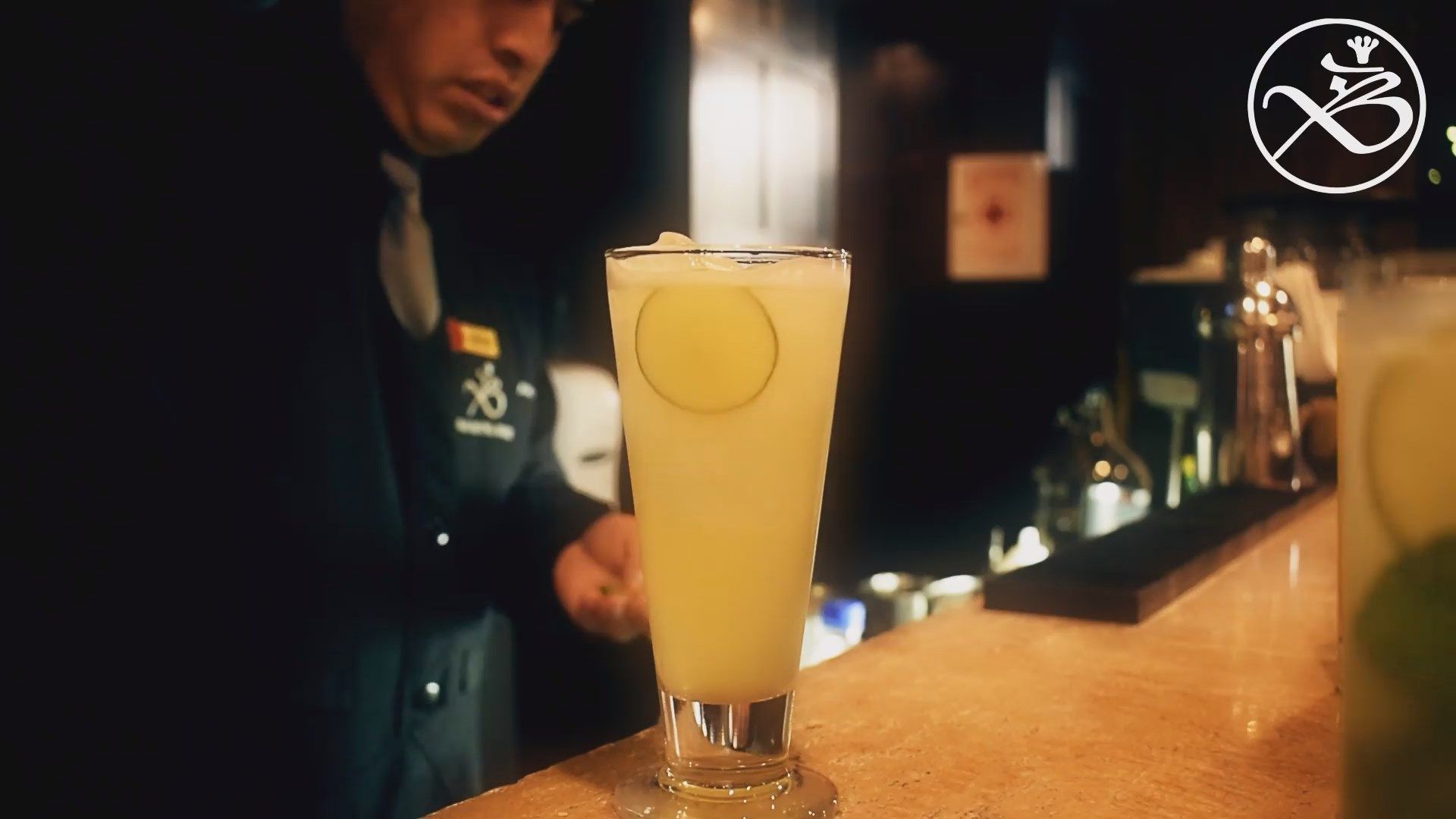 Cóctel amarillo en una copa alta sobre la barra, con un barman al fondo.