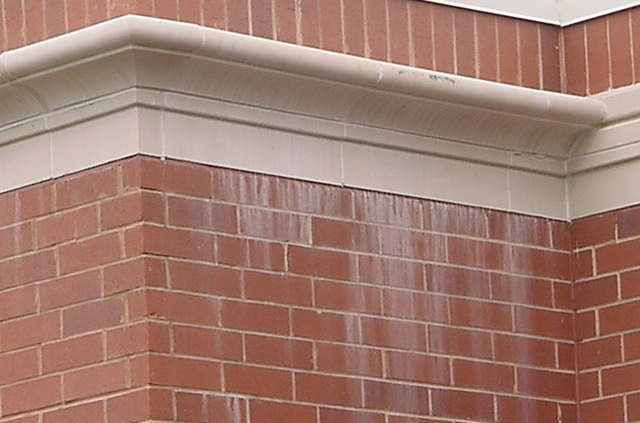 Brick building corner with light-colored trim at the top, water stains are visible on the brick.