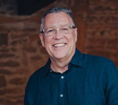 Smiling man in glasses and a navy shirt against a blurred brick wall background