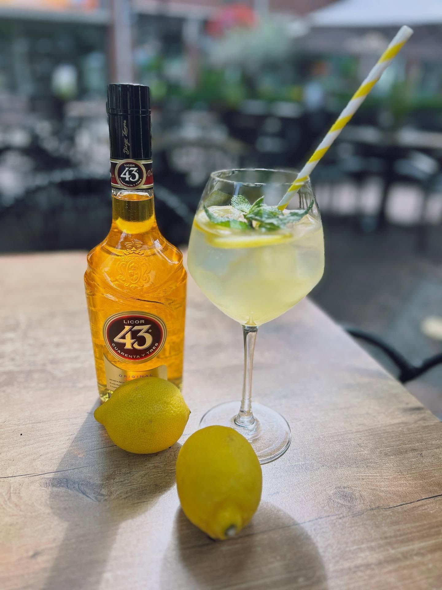 Een fles Licor 43 naast een gegarneerd cocktailglas met een rietje en twee verse citroenen op een houten buitentafel.