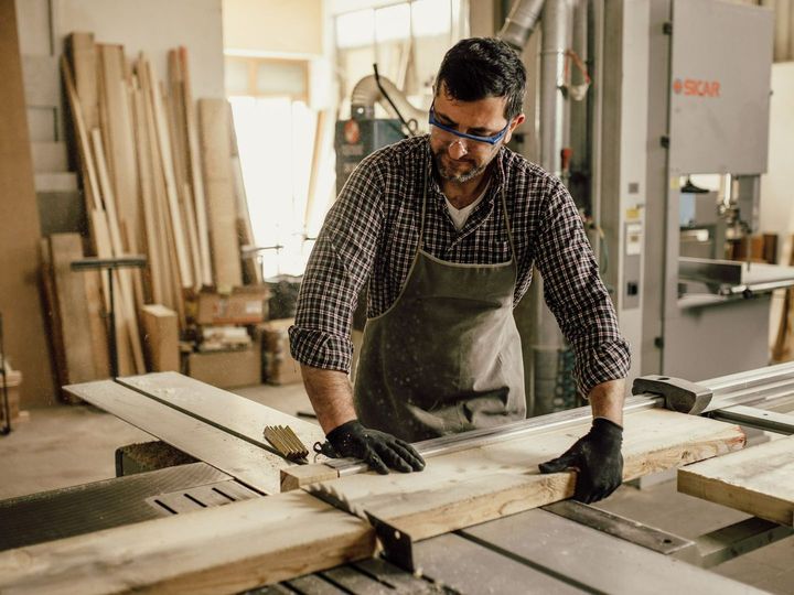 A man is cutting a piece of wood on a table saw in a workshop.