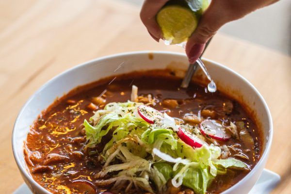 Hand squeezing lime over a bowl of red soup topped with shredded lettuce and radishes