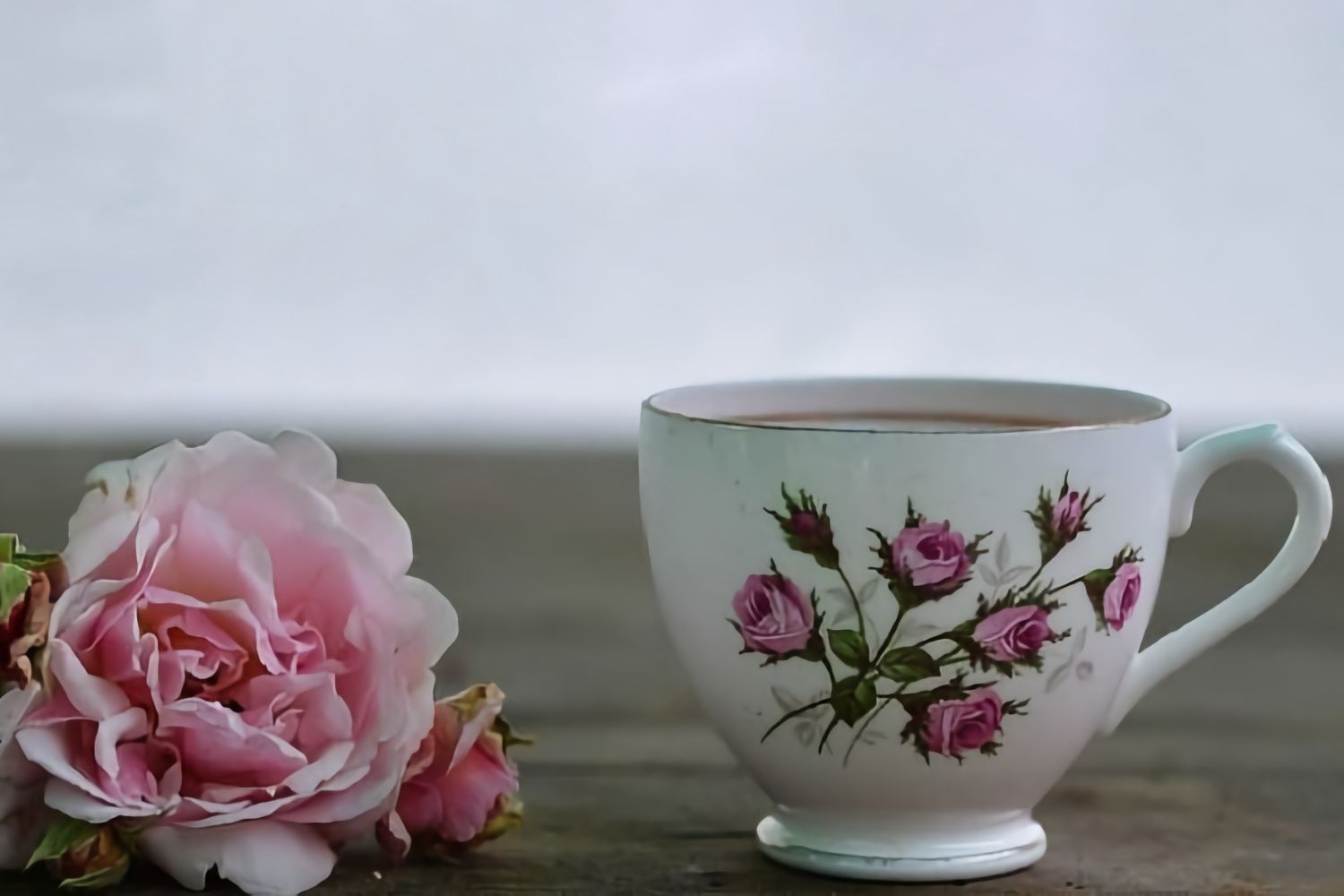 Pink rose beside a white teacup with floral design on a wooden surface