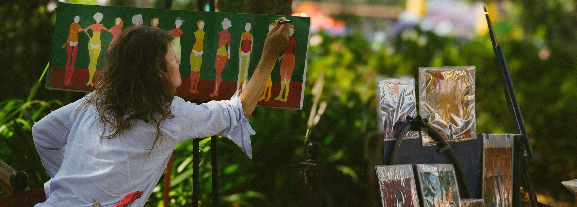 Artist painting colorful figures on an outdoor easel beside a display of framed artwork