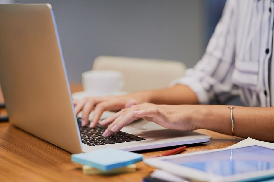 Person typing on a laptop at a wooden desk with papers and a smartphone nearby.
