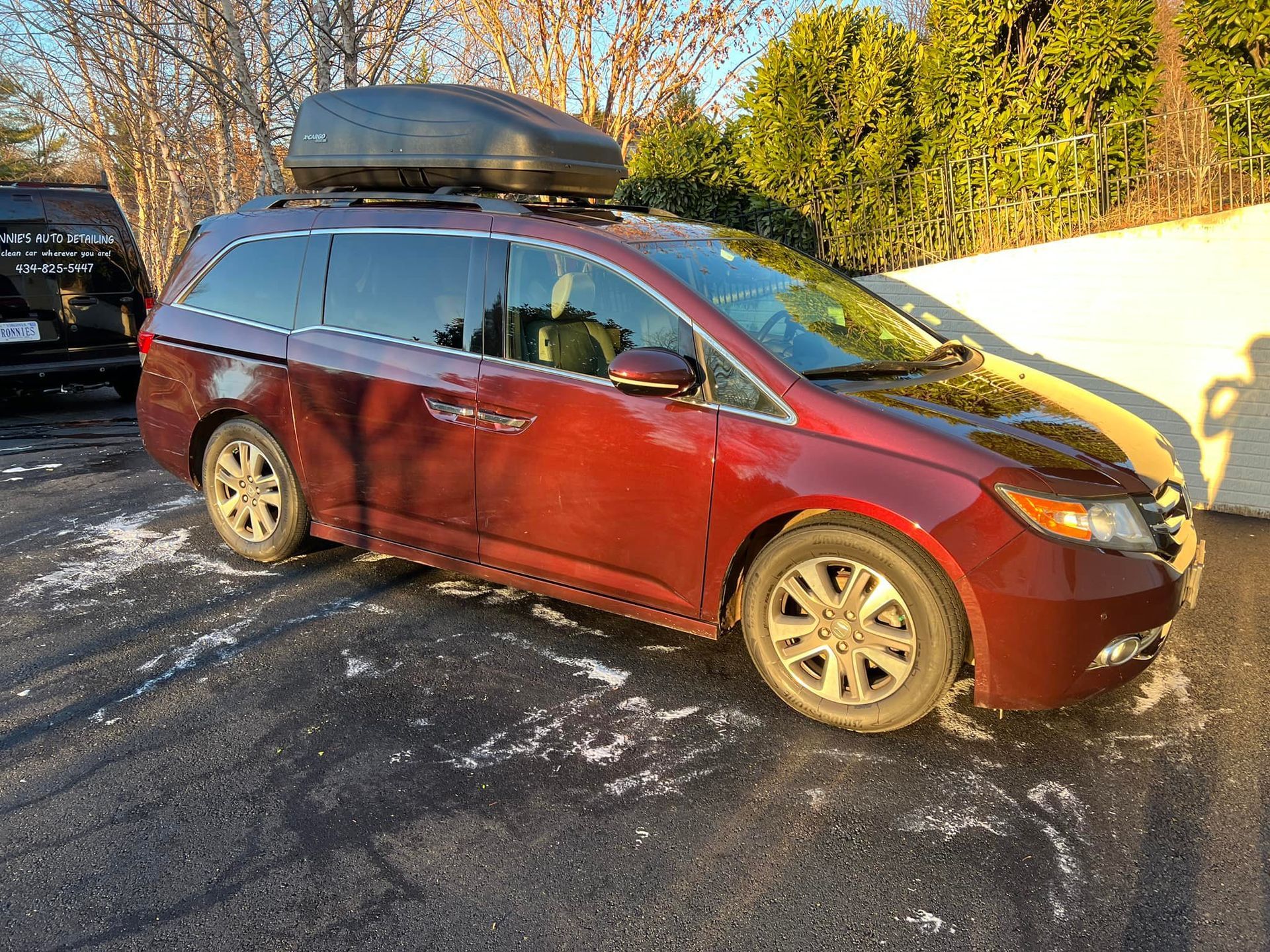 Red minivan with roof cargo box parked on a snowy driveway by a white fence and trees.