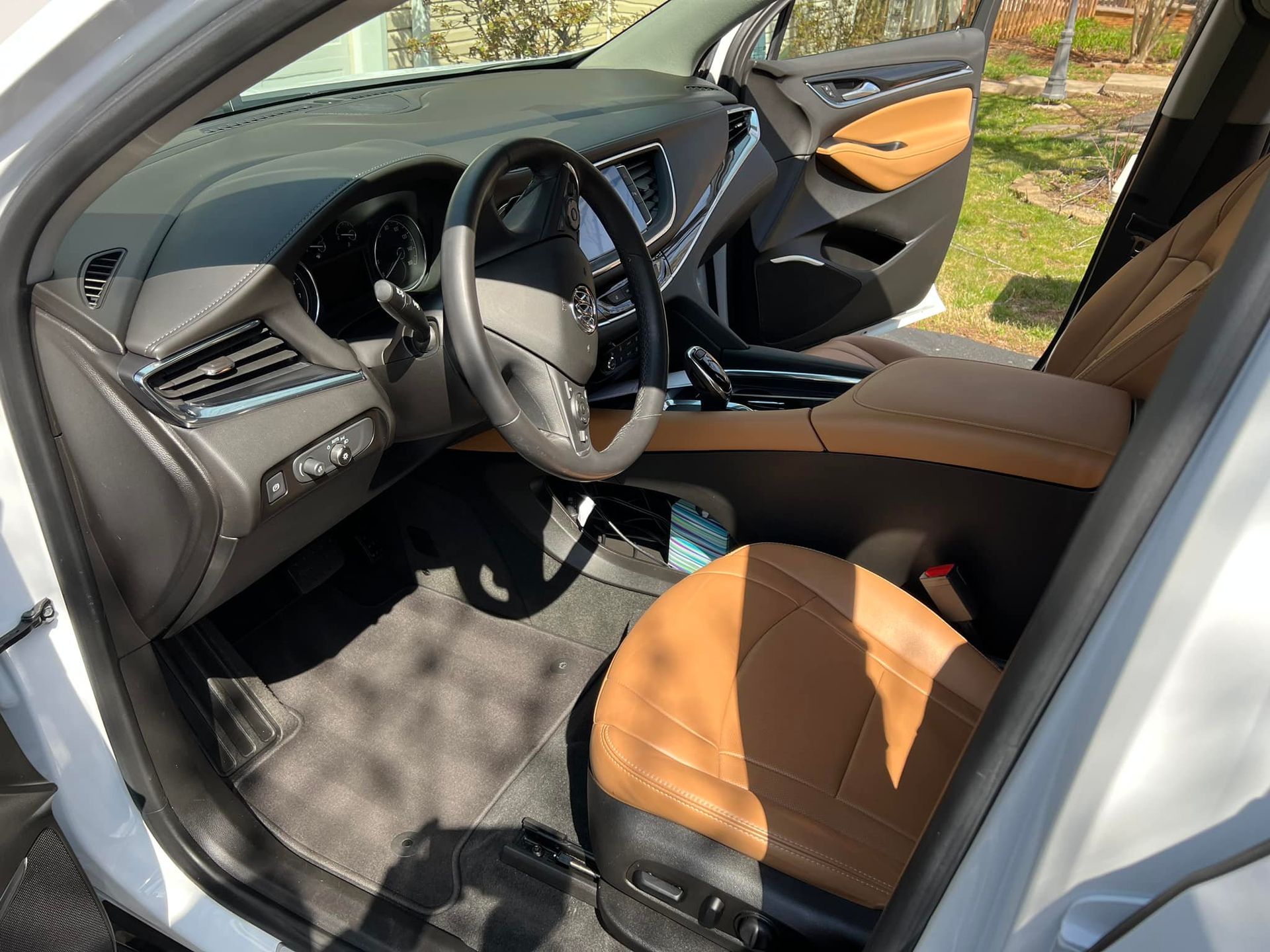 Car interior with black dashboard, tan seats, and open front passenger door beside a wooded area