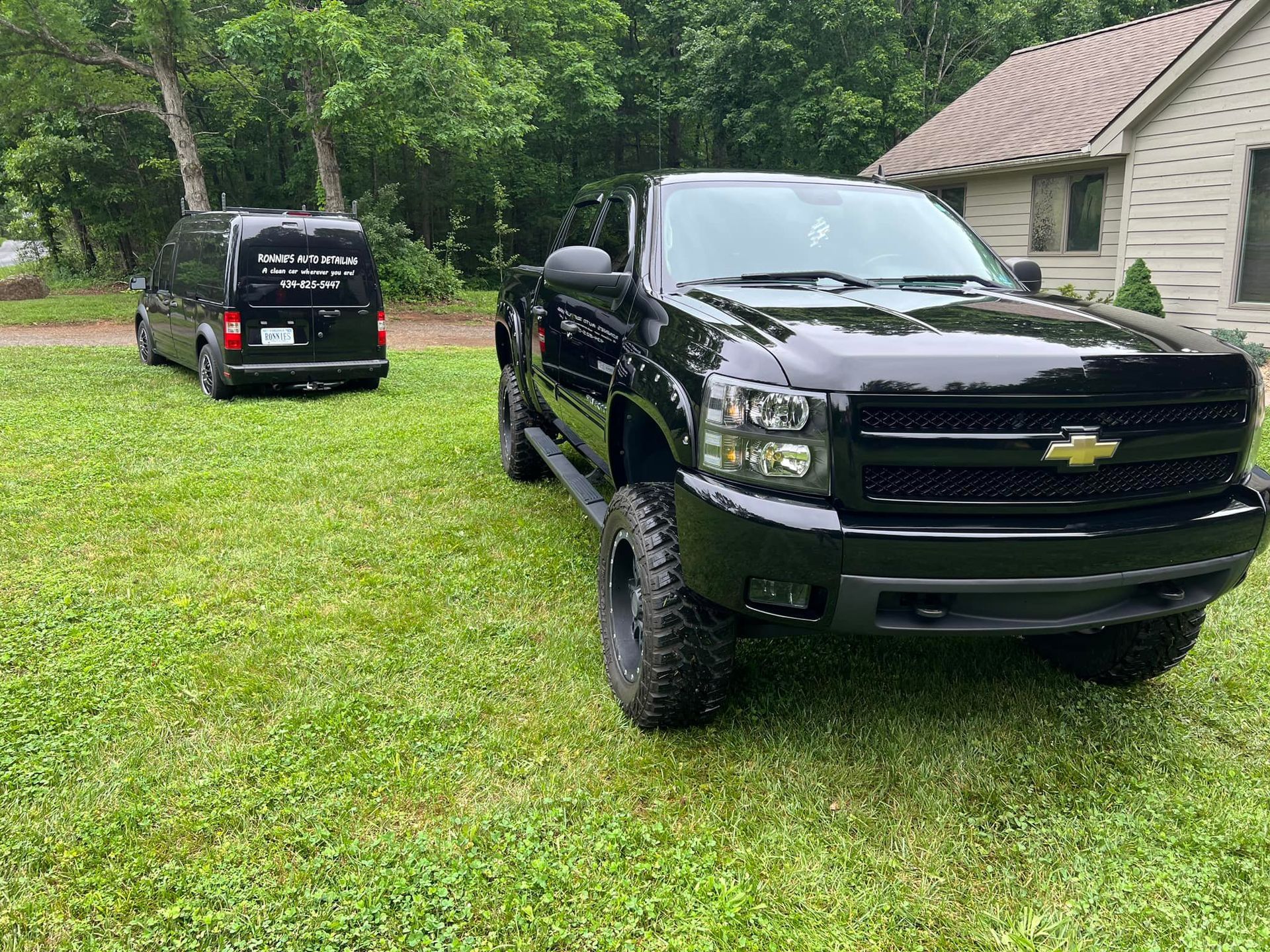 Black Chevrolet pickup and black van parked on grass in a wooded yard near a house.