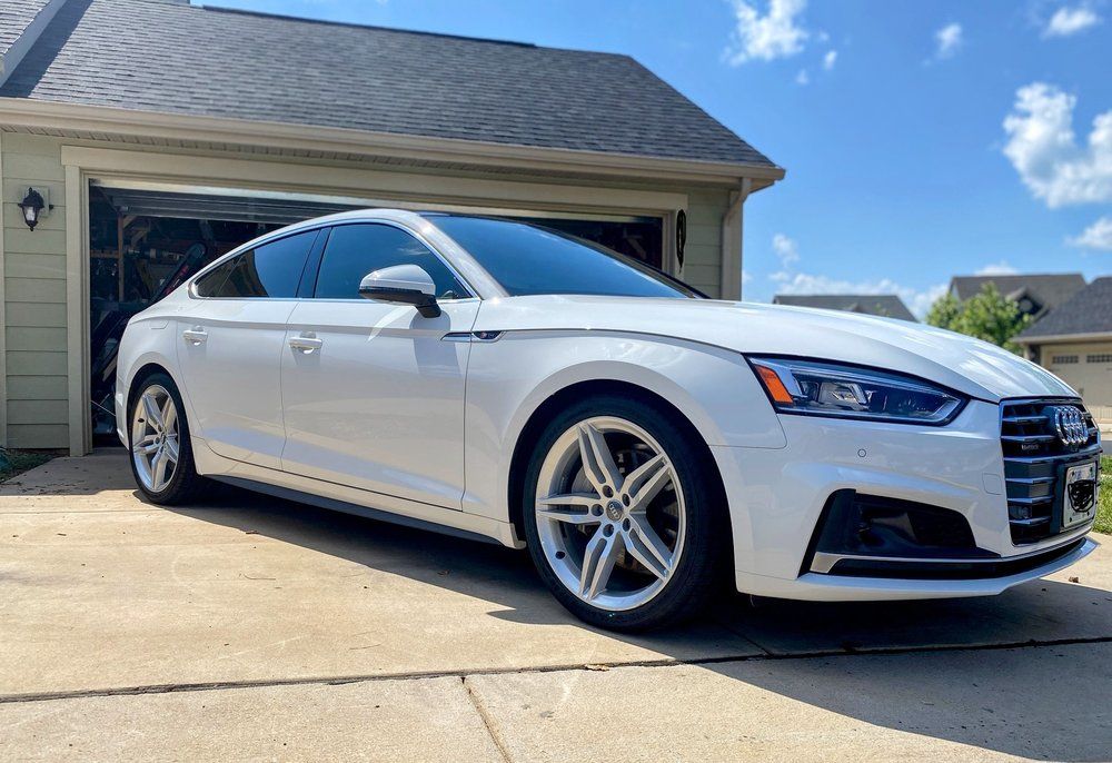 White sedan parked in a driveway beside a suburban house under a blue sky