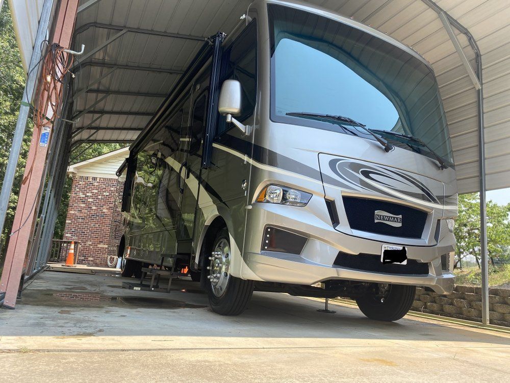Black and silver RV parked under a metal carport beside a brick building