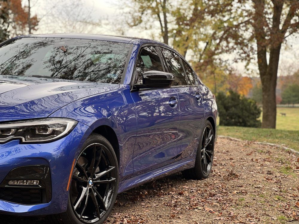 Blue sedan parked on a leaf-covered road beside trees in autumn