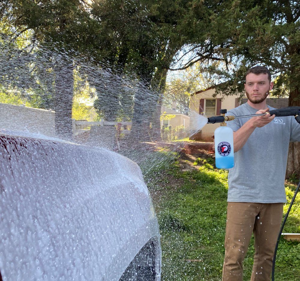 Man spraying a white car with a pressure washer in a sunny yard