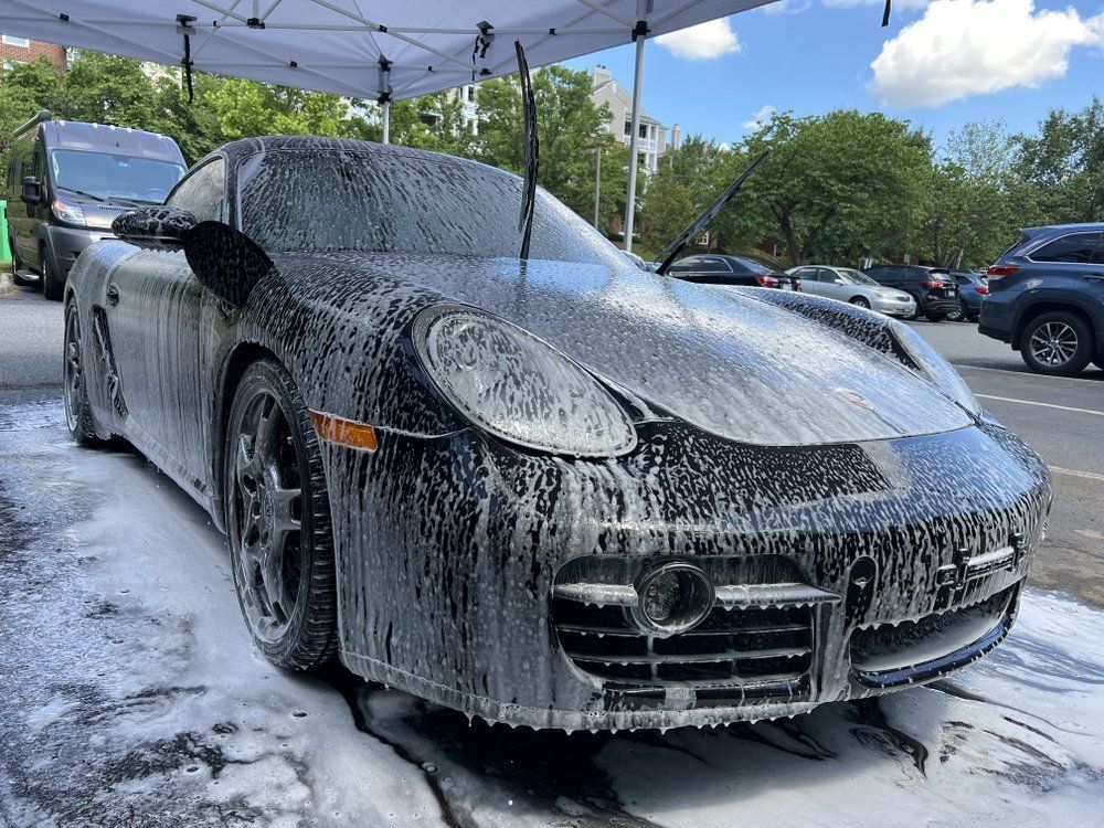Black sports car being hand-washed under a canopy, covered in thick soap suds on a sunny day.