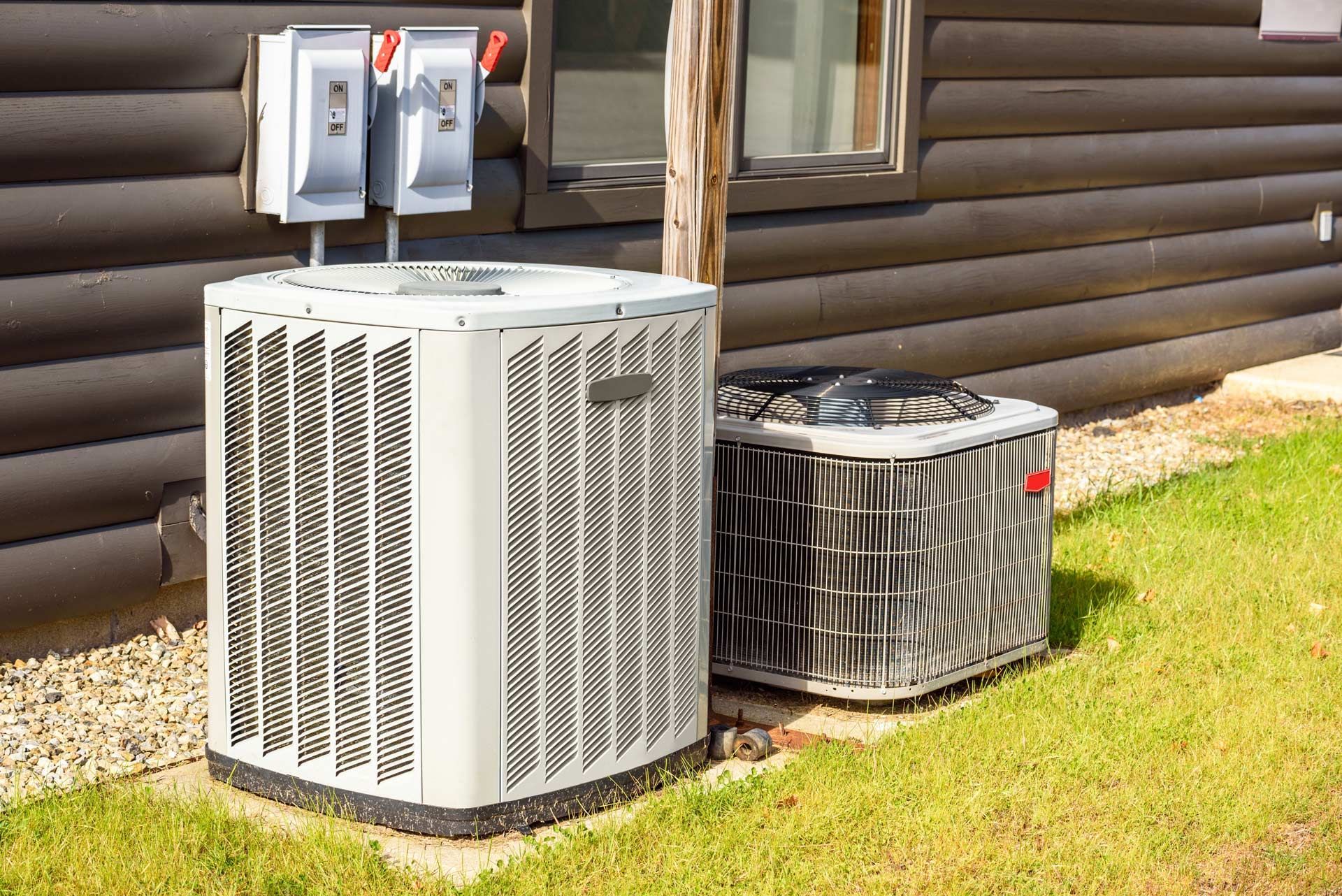 Four gray air conditioning units lined up outside a building with greenery.