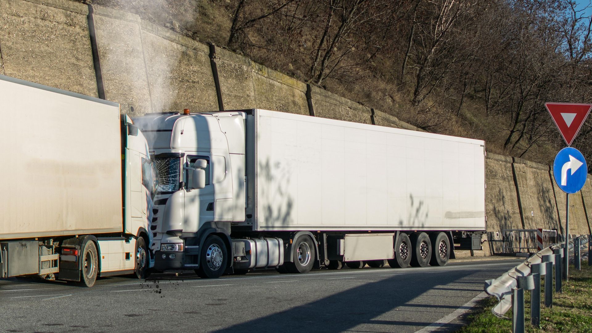 Two white semi-trucks parked along a roadside beside a stone wall and yield sign