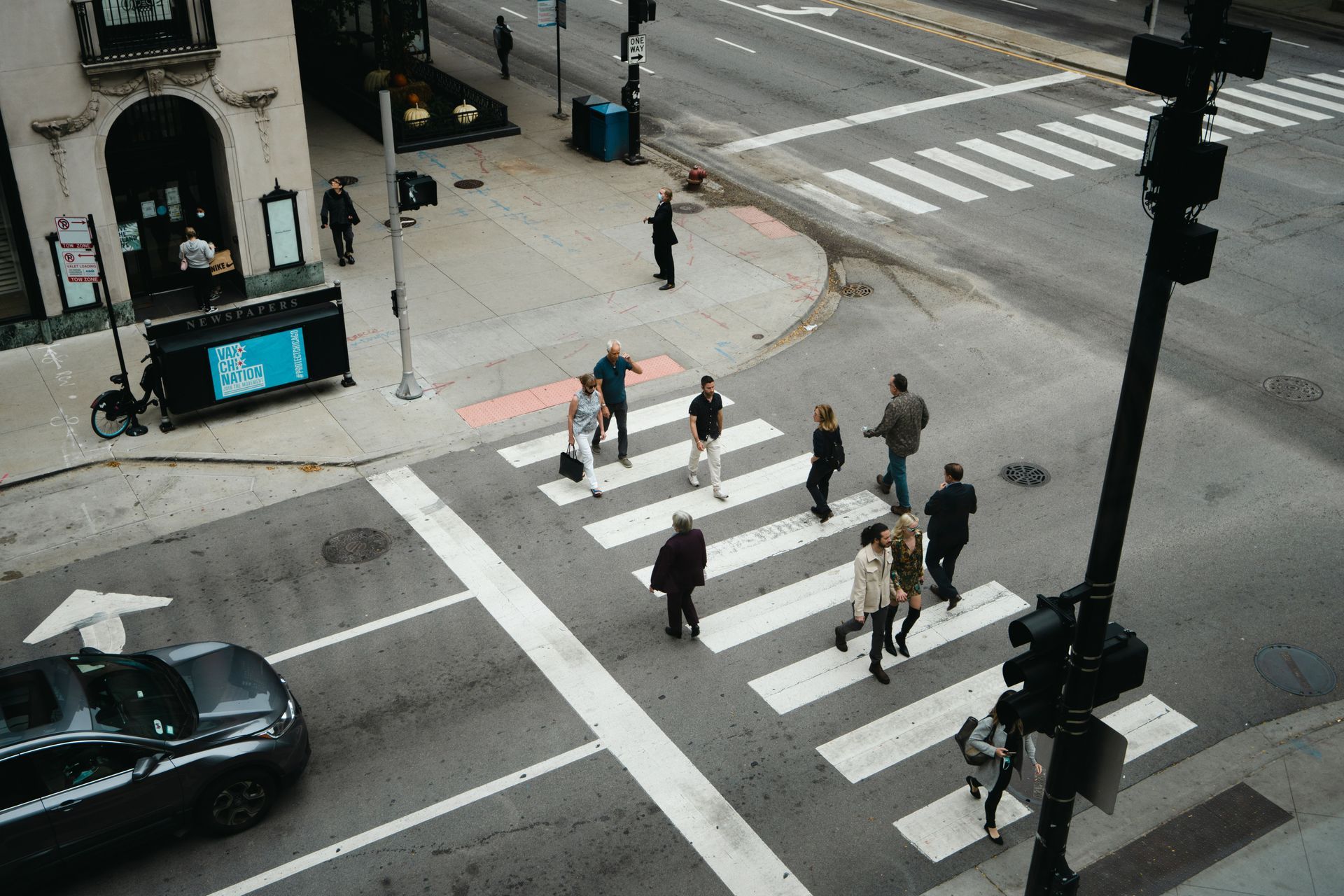 Pedestrians crossing a city intersection on a marked crosswalk near parked cars and traffic lights