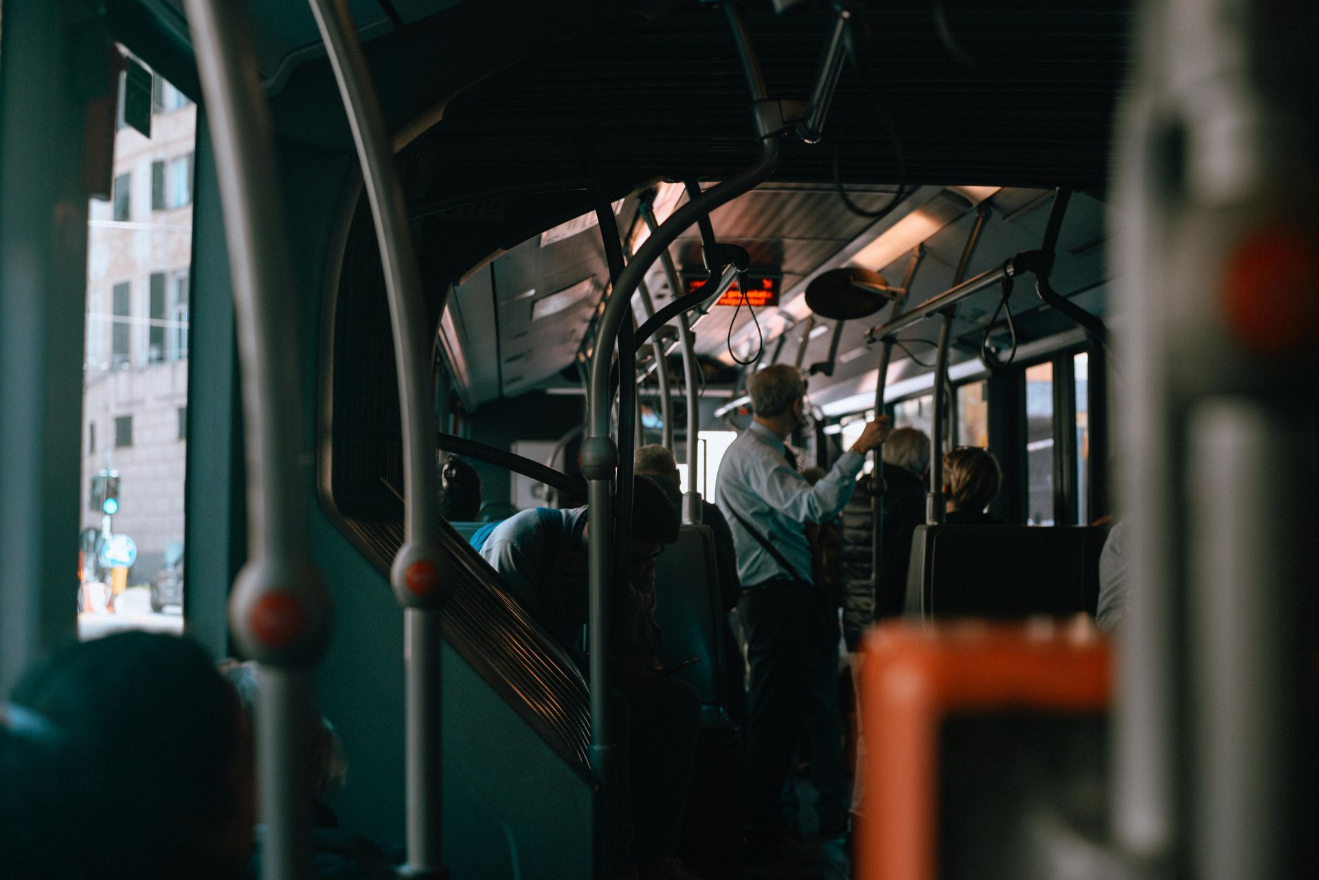 A person stands and holds a handrail inside a dimly lit bus, with other passengers seated nearby.