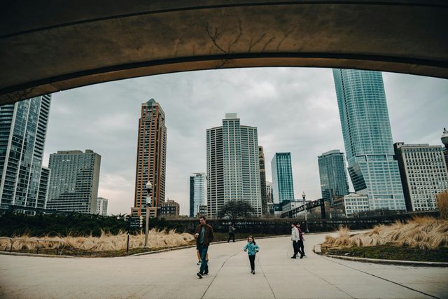 People walking on a wide path beneath an overpass, with tall city skyscrapers ahead.
