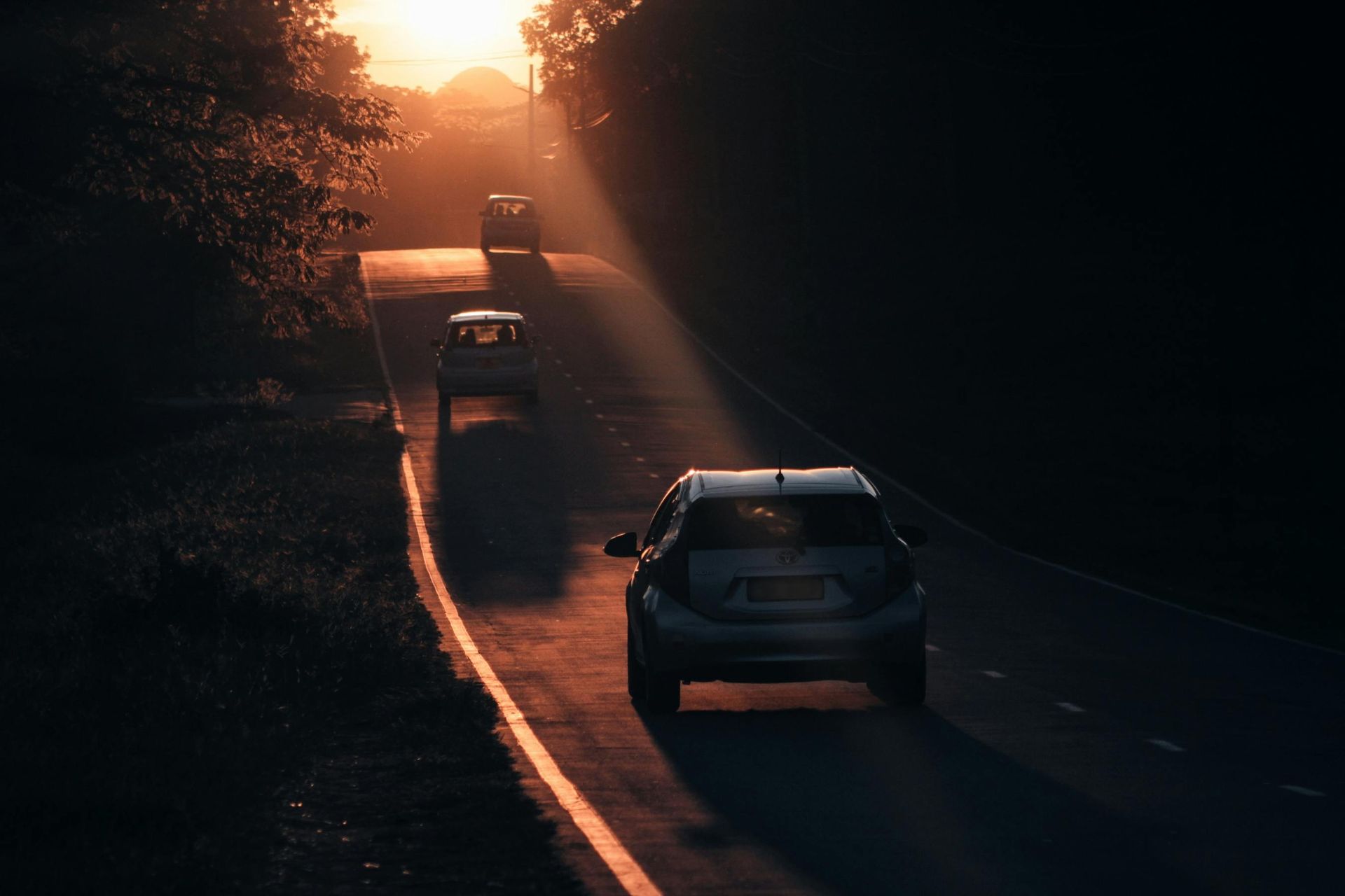 Three cars drive along a dark road toward a bright sunset on the horizon.