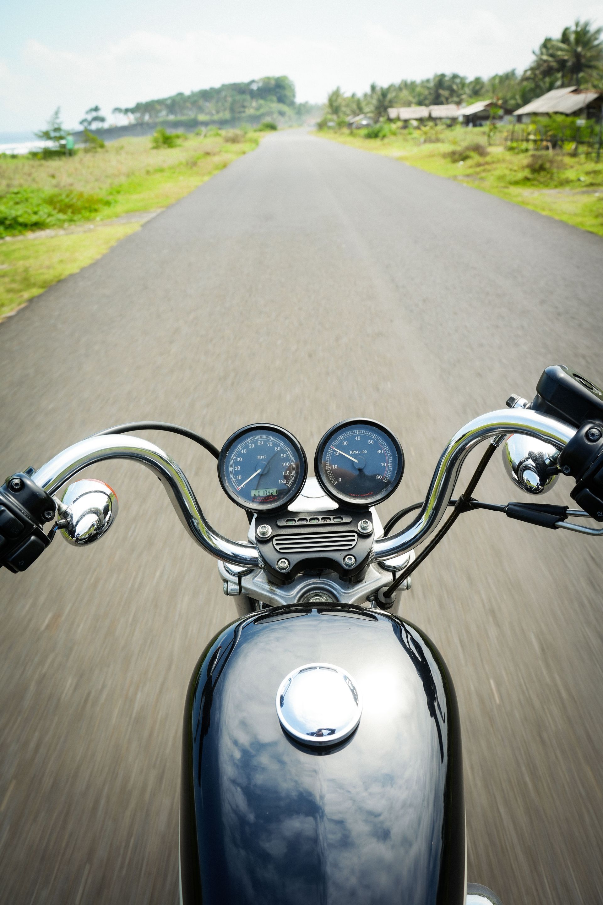 View from a motorcycle on a rural road, handlebars and gauges in foreground, greenery on both sides