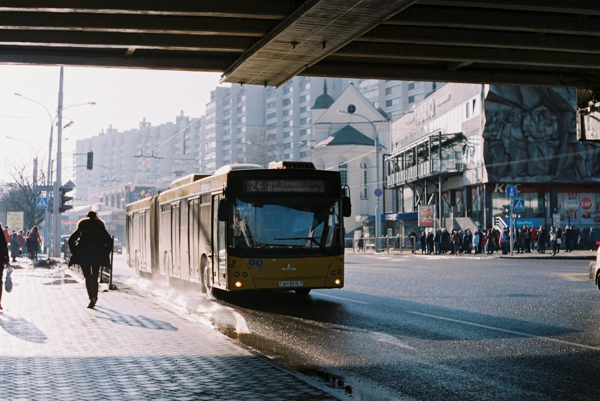 A yellow articulated bus travels down a city street under an overpass, with a pedestrian walking on the sidewalk.