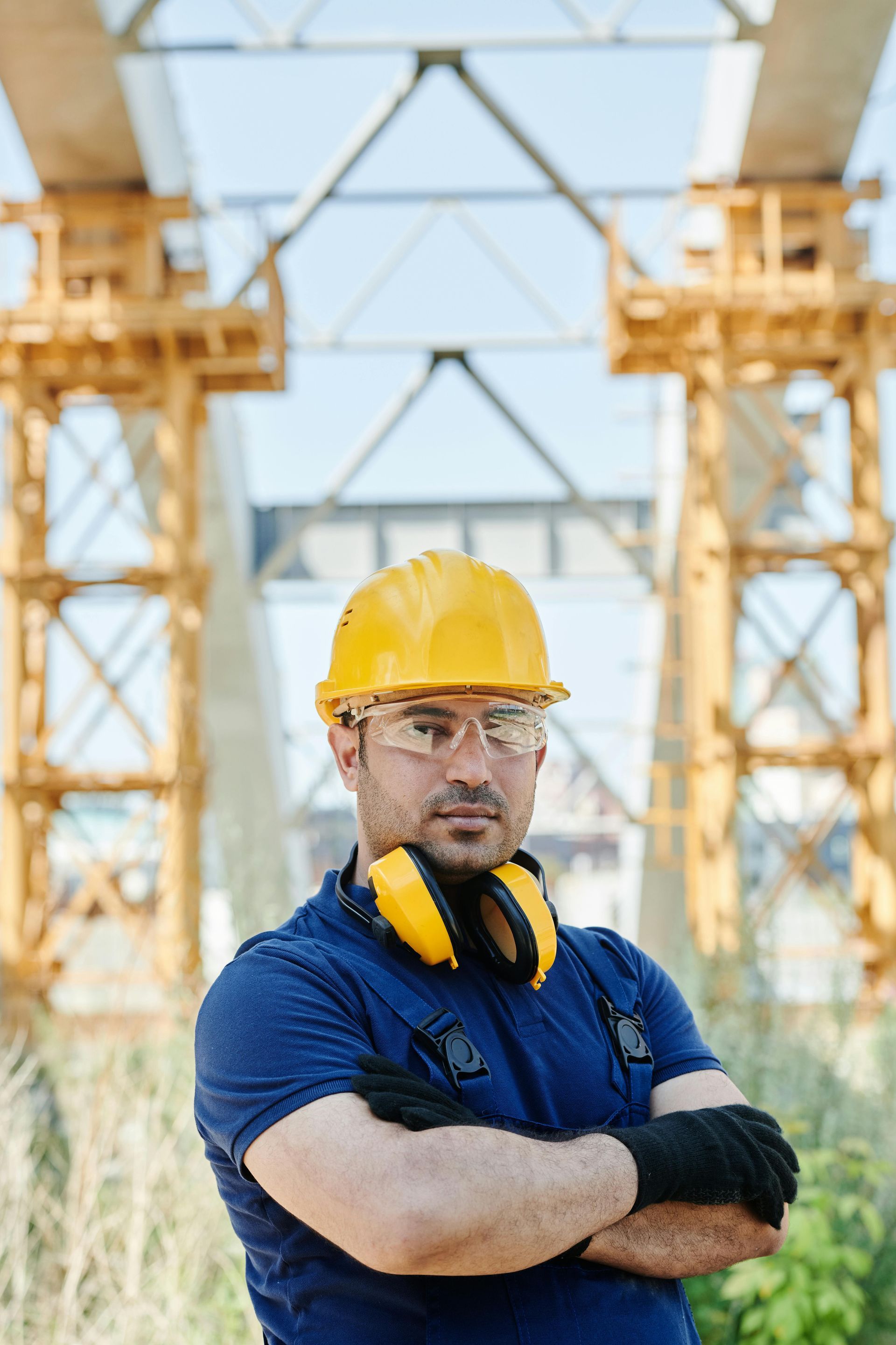A construction worker in a yellow hard hat, safety glasses, and ear protectors, standing with arms crossed at a bridge site.