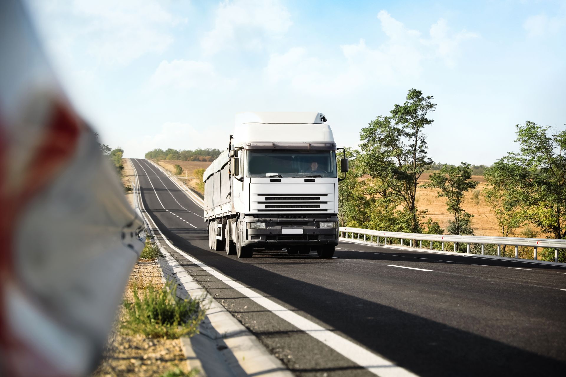 White semi-truck driving on a highway under a clear sky, with guardrails and trees along the road