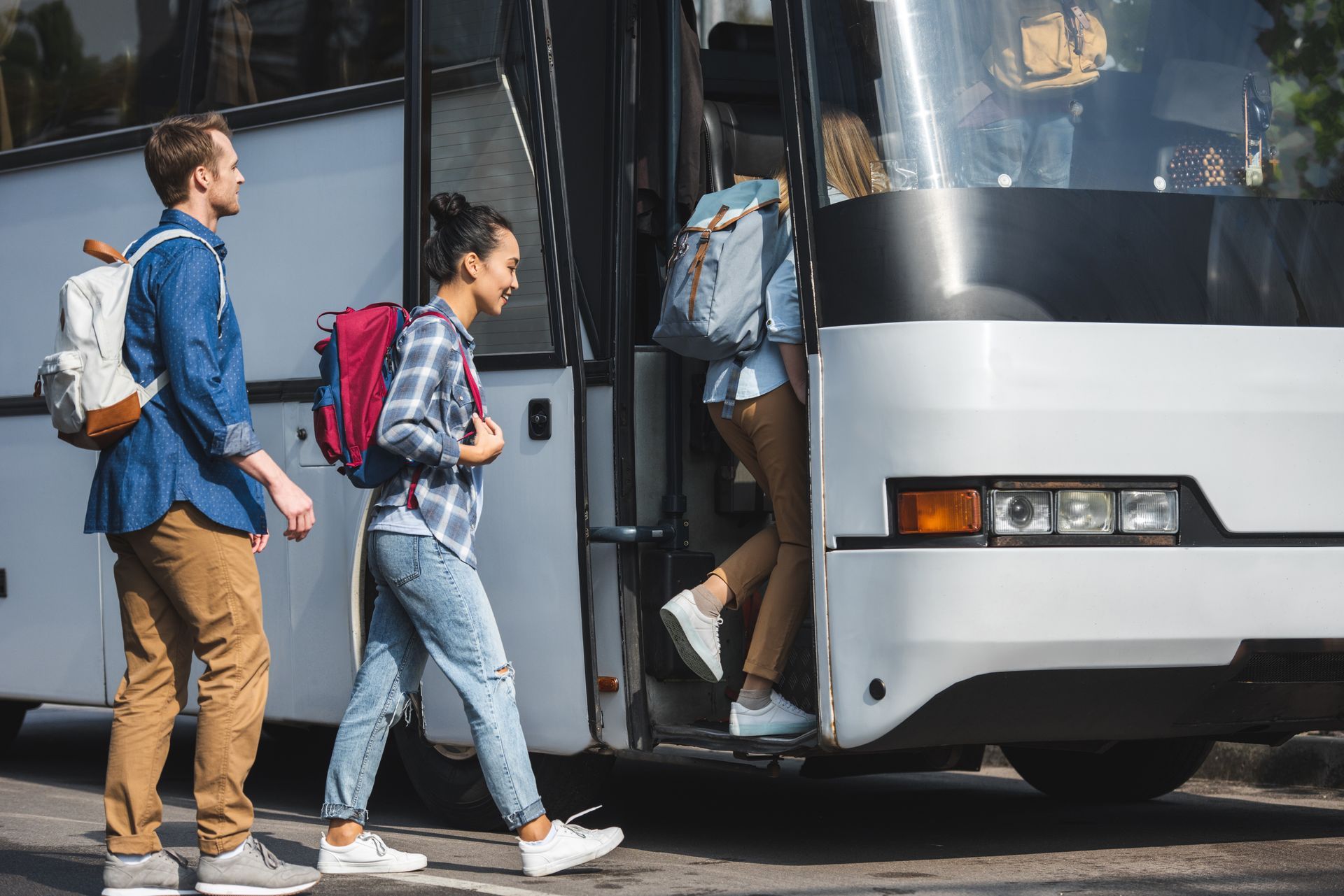 People boarding a white bus, carrying backpacks, in a parking lot