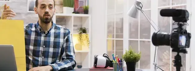 Man in a plaid shirt recording a video, with camera and microphone on a tripod.