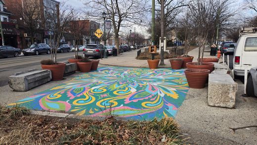 A streetscape with colorful mural, planters, benches, and parked cars along the sidewalk.