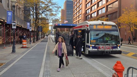 Bus at a stop in a city. People boarding. Woman in foreground. Buildings in background.