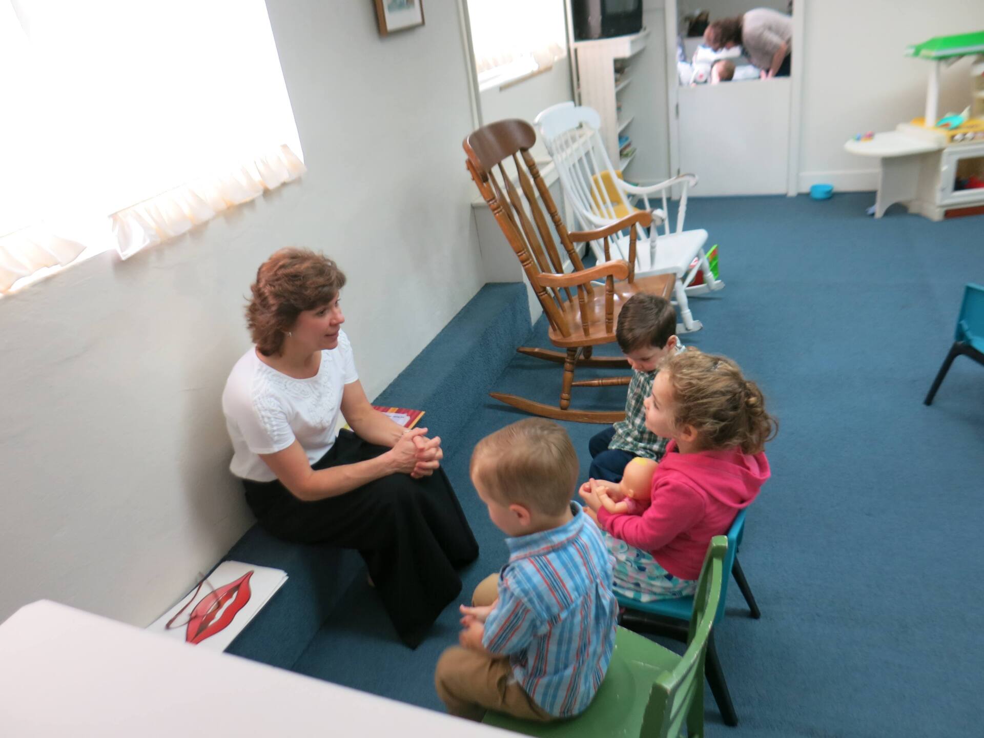 A woman is talking to a group of children in a room with rocking chairs
