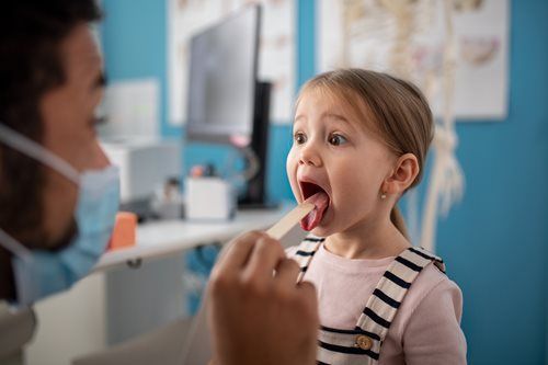 Child opening mouth for a throat exam in a clinic, with a doctor using a tongue depressor.